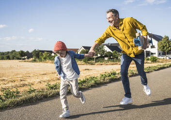 Cheerful man and boy holding hands and running on road - UUF30066