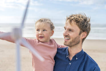 Lächelnder Mann mit Tochter, die ein Windradmodell am Strand hält - SBOF04015
