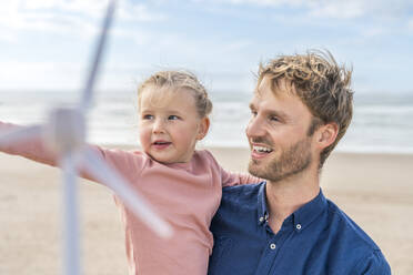 Smiling man with daughter holding wind turbine model at beach - SBOF04015