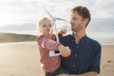 Father with daughter holding wind turbine model at beach on sunny day - SBOF04047