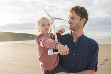 Vater mit Tochter, die an einem sonnigen Tag am Strand ein Modell einer Windkraftanlage halten - SBOF04047