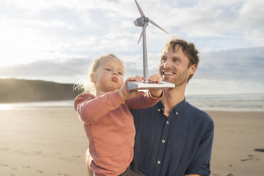 Smiling father with daughter holding wind turbine model under cloudy sky - SBOF04048