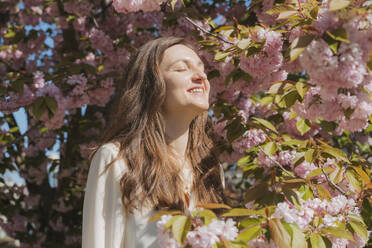 Glückliche Frau, die in der Nähe eines blühenden Kirschbaums an einem sonnigen Tag steht - OSF02092
