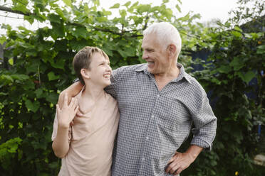 Happy grandfather standing with grandson in front of plants in garden - EYAF02764