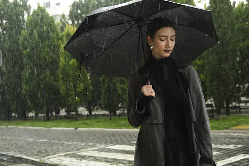 Young woman holding umbrella in rain on street - YHF00089