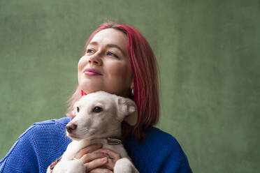 Lächelnde Frau mit gefärbtem Haar, die ihren Hund vor einer grünen Wand umarmt - OSF02103