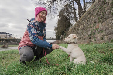 Lächelnde Frau mit Strickmütze beim Händeschütteln mit Hund auf Gras im Garten - OSF02106