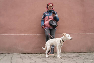 Lächelnde Frau, die mit ihrem Hund vor einer Wand steht und ihr Smartphone benutzt - OSF02110