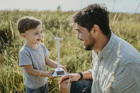 Lächelnder Sohn zeigt seinem Vater das Modell einer Windkraftanlage auf einem Feld - ANAF02073