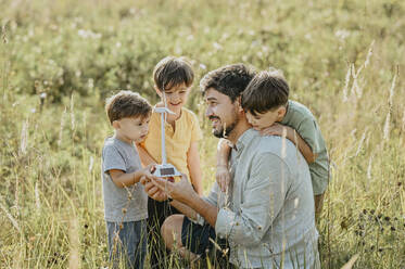Lächelnder Vater zeigt Kindern auf einem Feld das Modell einer Windkraftanlage - ANAF02075