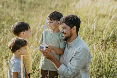 Glücklicher Vater zeigt Kindern auf einem Feld das Modell einer Windkraftanlage - ANAF02077