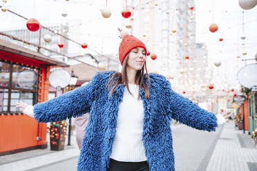 Smiling woman wearing Santa hat standing with arms outstretched on street - NLAF00089