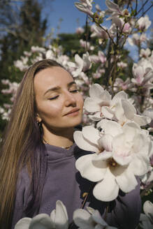 Frau mit geschlossenen Augen bei einer Magnolienblüte im Park an einem sonnigen Tag - YBF00190