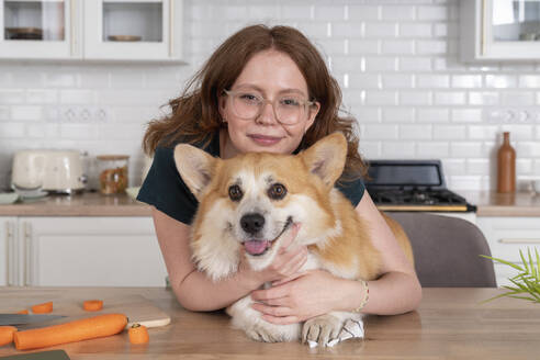 Lächelnde Frau umarmt Welsh Corgi Hund in der Küche zu Hause - VPIF08651