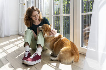 Smiling woman stroking Welsh Corgi dog sitting at home - VPIF08667