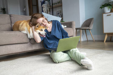 Frau, die einen Laptop benutzt und ihren Hund im Wohnzimmer umarmt - VPIF08692