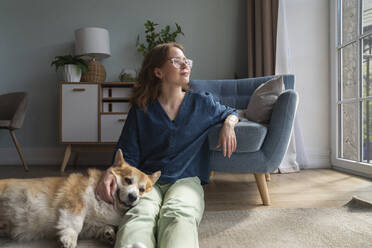 Thoughtful woman sitting with Welsh Corgi dog in living room - VPIF08700