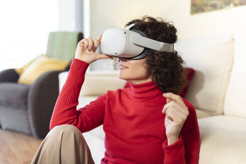 Smiling woman wearing virtual reality headset sitting at home - JPTF01259