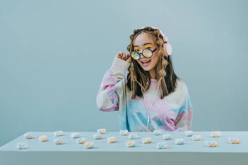 Glücklicher Junge mit Marshmallows auf einem Tisch und einer Kaleidoskop-Brille vor blauem Hintergrund - YTF01133