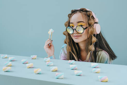 Junge Frau mit Kopfhörern und Kaleidoskopbrille isst Marshmallow im Studio - YTF01135