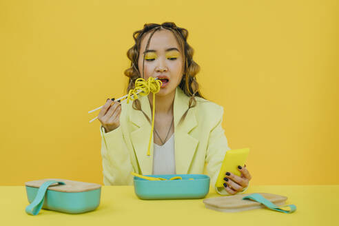 Junge Frau isst Nudeln und benutzt ein Mobiltelefon am Tisch im Studio - YTF01161