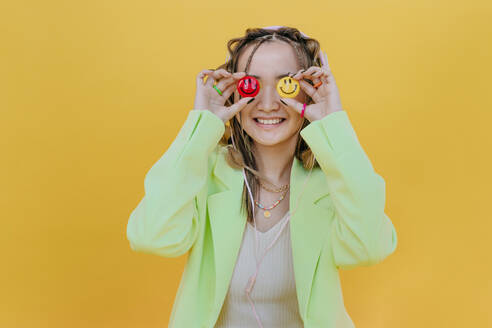 Lächelnde Frau, die ihre Augen mit Smileys vor gelbem Hintergrund bedeckt - YTF01171