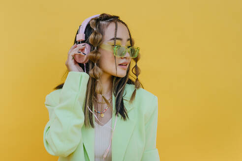 Young woman with sunglasses listening music on headphones against yellow background - YTF01179