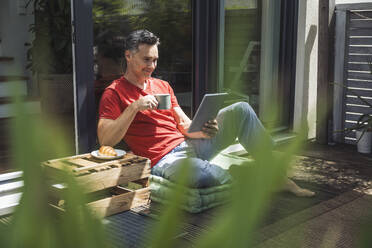 Man relaxing on balcony with digital tablet and mug in hands - UUF30091