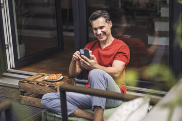 Man relaxing on balcony with smart phone and mug in hands - UUF30092