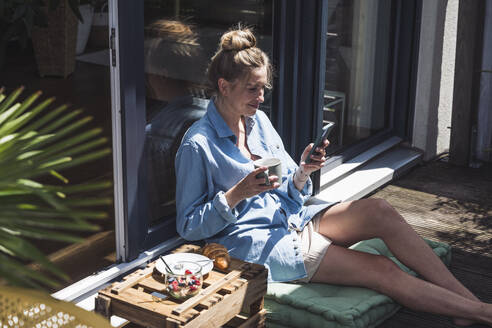 Porträt einer Frau, die sich auf einem Balkon mit einer Tasse und einem Smartphone in den Händen entspannt - UUF30118