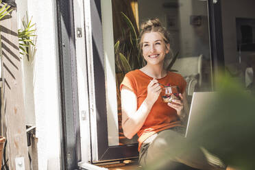 Portrait of woman sitting with laptop by open balcony eating dessert - UUF30127