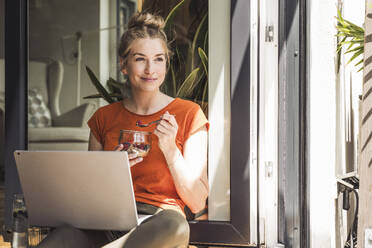 Porträt einer Frau, die mit einem Laptop vor einem offenen Balkon sitzt und ein Dessert isst - UUF30128
