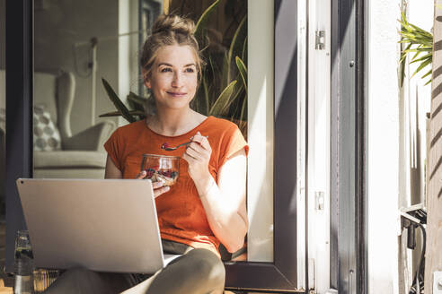 Portrait of woman sitting with laptop by open balcony eating dessert - UUF30128