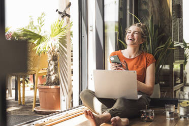 Portrait of woman sitting by open balcony laughing with smart phone in hands - UUF30132