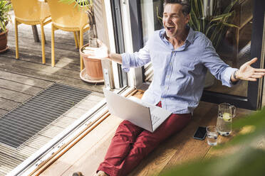 Porträt eines glücklichen Mannes mit Laptop auf einem offenen Balkon - UUF30138