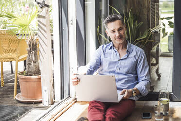 Portrait of man using laptop by open balcony - UUF30141