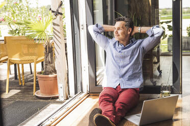 Portrait of man relaxing by open balcony with laptop - UUF30143