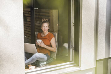Woman siting by window with laptop and mug - UUF30146