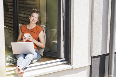 Woman siting by window with laptop and mug - UUF30147