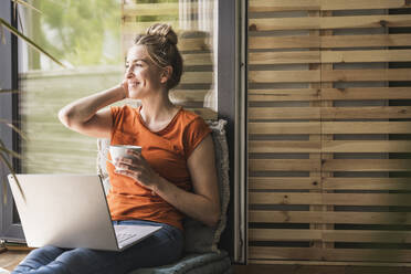Porträt einer Frau, die sich mit Laptop und Tasse auf dem Balkon entspannt - UUF30150