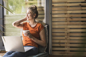 Porträt einer Frau, die sich mit Laptop und Tasse auf dem Balkon entspannt - UUF30150