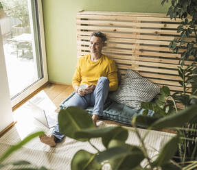 Portrait of man sitting on mattress in front of laptop - UUF30156