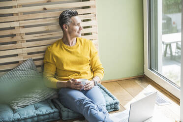 Porträt eines auf einer Matratze sitzenden Mannes vor einem Laptop - UUF30157