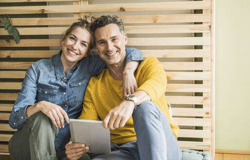 Portrait of couple sitting together in front of wooden bed frame leaning on wall - UUF30165