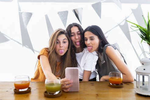 Friends sticking out tongue and taking selfie with smart phone at table - JPTF01273