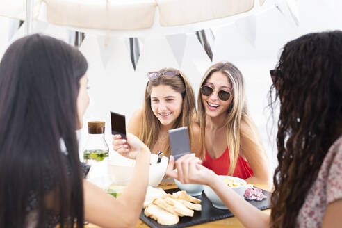 Smiling friends showing photographs at table - JPTF01277