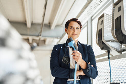 Confident businesswoman with electric plug at charging station - JOSEF20703