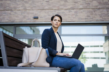 Confident businesswoman sitting with laptop on bench - JOSEF20714