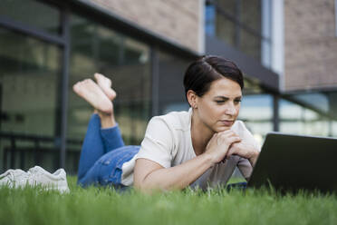 Businesswoman using laptop lying on grass at office park - JOSEF20721