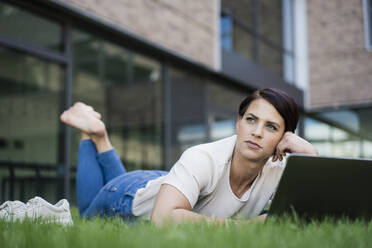 Contemplative businesswoman with laptop lying on grass - JOSEF20722